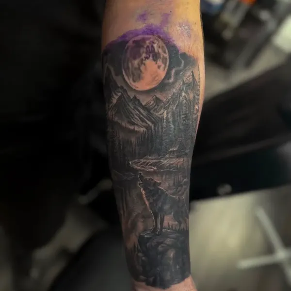 Wolf howling at a full moon over a mountain landscape, black and grey forearm tattoo by Kai at Shade Studio Peterborough